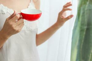 jovem mulher vestindo branco Vejo através pijamas em pé de janela dentro manhã e segurando copo do café para beber e Vejo lindo manhã cenário sozinho. conceito do bebendo café para estimular corpo. foto