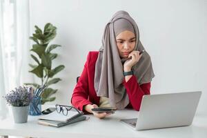 sério jovem mulher vestindo hijab sentado às escrivaninha trabalhando em Novo o negócio projeto sozinho dentro moderno escritório foto