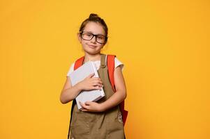 pequeno criança menina segurando livro, sorridente olhando às Câmera, vestido dentro casual roupas e na moda óculos. costas para escola foto