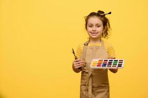 confiante criativo sorridente pequeno criança menina detém aguarela paleta, pintura escova e paleta faca, isolado em amarelo foto