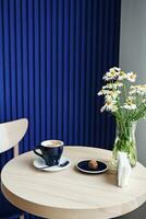 ainda vida. camomilas dentro uma vaso perto uma copo do café e uma delicioso doce Brigadeiro em uma de madeira mesa Próximo para a gravado azul muro. foto