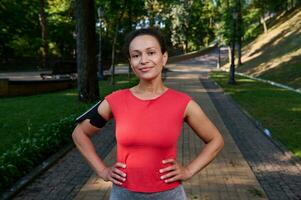 lindo muscular mulher com saudável estético corpo e brilhando pele, atleta dentro vermelho camiseta e Smartphone suporte posando com braços em cintura olhando às Câmera em pé em a cidade parque esteira foto