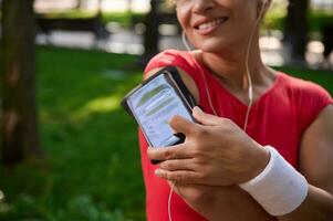 foco em Smartphone suporte em a braço do borrado atleta colocando dedo em a telefone e ajustando Móvel aplicativo e lista de reprodução durante manhã correr foto