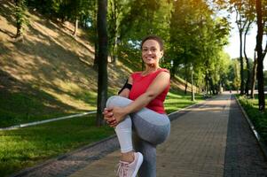 encantador jovem africano americano desportista, determinado atleta alongamento dela pernas, aquecimento acima dela corpo antes corpo peso exercite-se ao ar livre dentro cidade parque e sorrisos olhando às Câmera foto