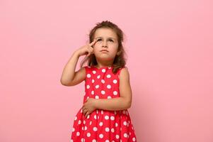 fofa adorável pré escola garota, 3-4 anos velho, parece pensativamente acima, segurando dela dedo para dela têmpora, posando em uma Rosa fundo com cópia de espaço. retrato do uma pensativo bebê menina foto