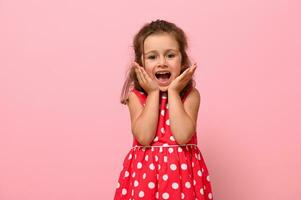 fechar-se retrato do uma linda bebê menina dentro uma Rosa vestir com polca pontos poses às a Câmera, em pé contra uma Rosa fundo e segurando dela queixo com dela mãos. foto