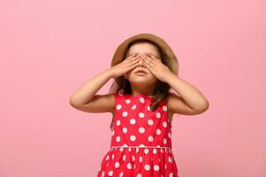 confiante retrato do linda bebê menina vestido dentro verão polca pontos vestir e Palha chapéu , cobertura dela olhos com dela mãos, posando sobre Rosa fundo com cópia de espaço para publicidade foto