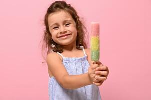 engraçado alegre pequeno menina sorrisos mostrando para a Câmera uma delicioso doce congeladas suco, picolé, gelo creme em bastão dentro dela mãos. verão conceito, Rosa fundo, cópia de espaço foto