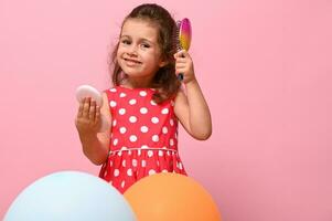 linda adorável bebê menina dentro à moda Rosa vestir, olhando às Câmera, em pé atrás multicolorido balões, pentear dela encaracolado cabelo , posando com pequeno Cosmético espelho dentro mãos. cópia de espaço foto