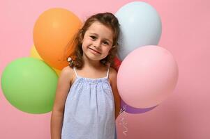 lindo pequeno aniversário 4 anos menina fofa sorrisos segurando multicolorido balões atrás dela voltar, isolado sobre Rosa fundo, cópia de espaço. retrato do linda pequeno menina para anúncio. foto