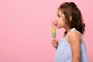 fofa bebê menina desfrutando delicioso colorida gelo creme picolé em bastão isolado em Rosa fundo com cópia de espaço. pequeno bebê comendo vegano colori sorvete foto