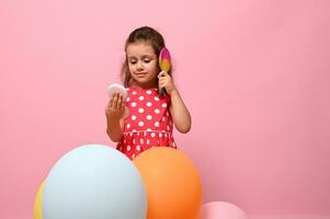 isolado retrato em Rosa fundo do uma linda bebê menina dentro à moda Rosa vestir, em pé atrás multicolorido balões, pentear dela encaracolado cabelo , olhando às dela reflexão dentro pequeno Cosmético espelho. foto