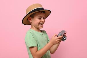 feliz criança dentro verão equipamento segurando uma retro vintage Câmera e fofinho sorrisos com cheio de dentes sorrir posando sobre Rosa fundo com cópia de espaço. turismo conceitos foto