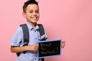 adorável estudante com mochila detém quadro-negro com giz letras ,voltar para escola, e fofa sorrisos para Câmera, isolado sobre Rosa fundo com espaço para texto foto