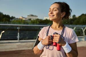 feliz confiante desportista com uma saltando corda, posando com fechadas olhos, em pé em a moderno vidro cidade ponte, desfrutando cedo ensolarado manhã cardio exercite-se ar livre. cardio exercite-se conceito foto