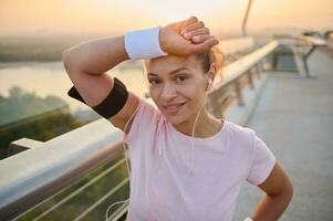 lindo hispânico desportista, Exausta fêmea atleta lenços suor a partir de dela testa com Terry pulseiras, sorrisos olhando às Câmera em pé em a cidade ponte, em repouso depois de uma manhã correr às alvorecer. foto