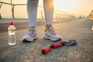 recortado imagem do atleta pernas vestindo Rosa tênis e em pé em a asfalto esteira Próximo para uma deitado baixa pulando corda e água garrafa contra a fundo do uma lindo nascer do sol em a ponte foto