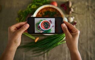 a sobrecarga Visão do mãos segurando Smartphone e fotografando uma Panela com recentemente cozinhou ucraniano tradicional sopa- borscht com vegetação deitado em uma linho toalha de mesa. Móvel afiar dentro viver Visão modo foto