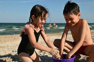 irmão e irmã, amigos, garoto e menina o preenchimento plástico brinquedo balde com molhado areia e construção arenoso figuras. feliz crianças jogando em a de praia às mar fundo, desfrutando verão Férias foto