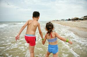 traseiro Visão do adorável crianças, Garoto e garota, irmão e irmã, caminhando mão dentro mão em a Beira Mar. verão Férias, Férias e Férias, felicidade e bem estar conceito foto