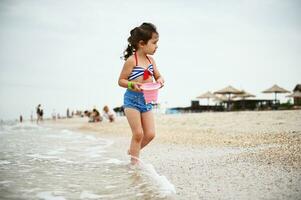 adorável bebê menina dentro colorida roupa de banho caminhando ao longo a Beira Mar com uma Rosa balde dentro dela mãos, cheio do mar água. lindo criança desfrutando verão Férias às mar, banhos de sol e mar jogos. foto