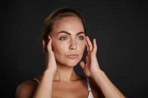 fechar-se retrato do a atraente caucasiano mulher com fresco brilhando pele segurando mãos em templos, fazendo massagem movimentos em dela face. anti-envelhecimento conceito, alisamento, rejuvenescedor beleza tratamento foto