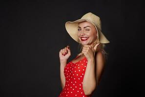 atraente deslumbrante Loiras mulher dentro verão Palha chapéu e vermelho roupa de banho piscando e sorridente com lindo branco cheio de dentes sorriso, mostrando polegar acima às Câmera, posando contra Preto fundo com cópia de espaço foto