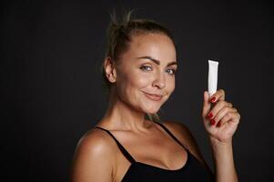 lindo mulher com bronzeada pele segurando uma tubo do anti rugas alisamento anti inchaço debaixo olhos creme, sorrisos olhando às Câmera, isolado sobre Preto fundo com cópia de de Anúncios espaço foto