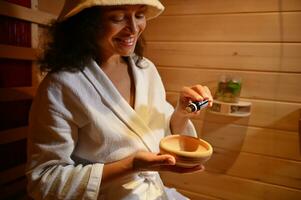 sorridente mulher derramando poucos gotas do manjericão essencial óleo para dentro de madeira argamassa enquanto relaxante dentro de madeira sauna foto