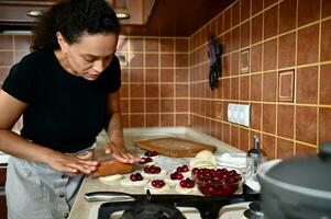 jovem mulher, chefe de cozinha pastelaria, rolos Fora massa com uma rolando PIN dentro volta formas em a cozinha bancada, prepara caseiro delicioso cereja dumplings foto