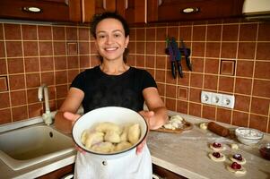 uma lindo misturado raça mulher chefe de cozinha pastelaria carrinhos contra a fundo do casa cozinha e sorrisos fofinho às a Câmera alongamento dela mãos com uma tigela do caseiro recentemente fervido dumplings foto