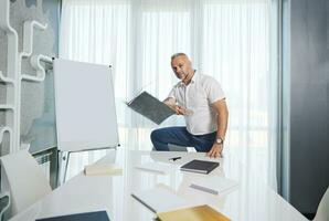 bonito meio envelhecido europeu homem, bem sucedido empreendedor, psicólogo, em pé de mesa perto uma giro gráfico com em branco espaço para texto, jogando uma livro , com confiança olhando às Câmera foto