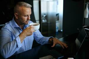 encantador meio envelhecido europeu homem relaxante, bebendo café enquanto trabalhando em computador portátil, sentado às mesa às casa escritório durante bloqueio foto