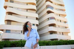 o negócio mulher dentro casual camisa, sorridente, falando em Móvel telefone, em pé contra moderno Alto subir edifícios fundo foto