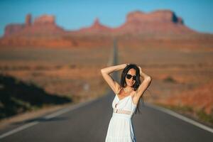 feliz jovem mulher dentro branco vestir em a famoso estrada para monumento vale dentro Utah dentro dela viagem dentro EUA. surpreendente Visão do a monumento vale. foto