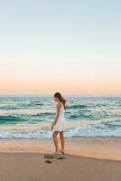 adorável adolescente menina dentro branco em a Beira Mar durante dela de praia verão período de férias com surpreendente céu em fundo foto
