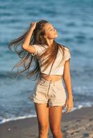 adorável jovem menina com lindo grandes cabelo apreciar tropical de praia período de férias. a menina em a Beira Mar às pôr do sol foto