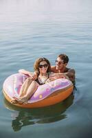 feliz jovem casal tomando banho dentro a mar em inflável flutuador dentro rosquinha forma foto