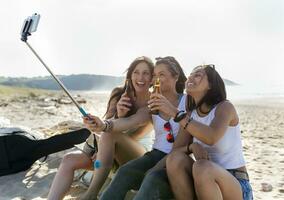 feliz fêmea amigos com Cerveja garrafas levando uma selfie em a de praia foto