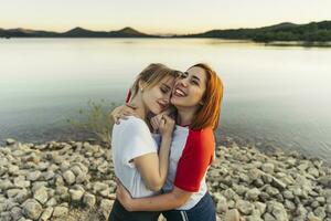 feliz mulher abraçando namorada enquanto em pé de lago durante pôr do sol foto