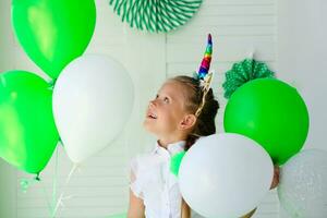 pequeno menina com uma unicórnio aro em dela cabeça contra a fundo do verde balões. aniversário para crianças. st. patrick's dia celebração foto