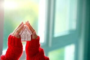 mulheres é mãos dentro uma vermelho suéter estão segurando uma Natal brinquedo dentro a forma do uma casa. fique lar. foto