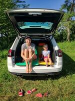 jovem mãe com dela pequeno filha sentado descalço dentro uma carro tronco dentro campo. feliz família indo para gastar a noite dentro a branco estação vagão dentro natureza. conceito do viajando, descansar e estrada viagem piquenique foto