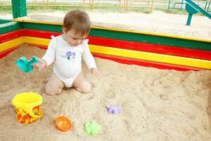 bebê menina para a primeiro Tempo dentro a caixa de areia parece às a brinquedos dentro confusão foto