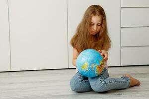 menina sentado em a chão cuidadosamente examina a globo dentro dela mãos foto