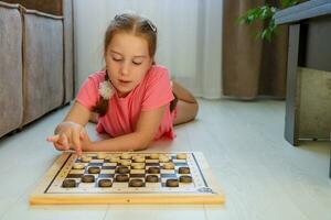 engraçado menina deitado em a chão às casa jogando jogo de damas foto