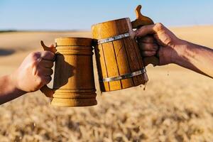 mãos com de madeira Cerveja tilintar canecas em a fundo do uma campo do trigo foto