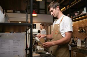 retrato do uma jovem masculino barista dentro avental usando Forma de pagamento máquina dentro cafeteria foto