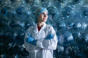 retrato do fêmea cientista com braços cruzado dentro bebendo água fábrica, ela estava em pé dentro frente do uma plástico garrafa. foto