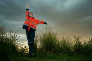 jovem mulher dentro engenheiro uniforme e Alto visibilidade com elevado braços em pé em gramíneo campo às pôr do sol, a conceito do relaxar Tempo depois de trabalhos foto